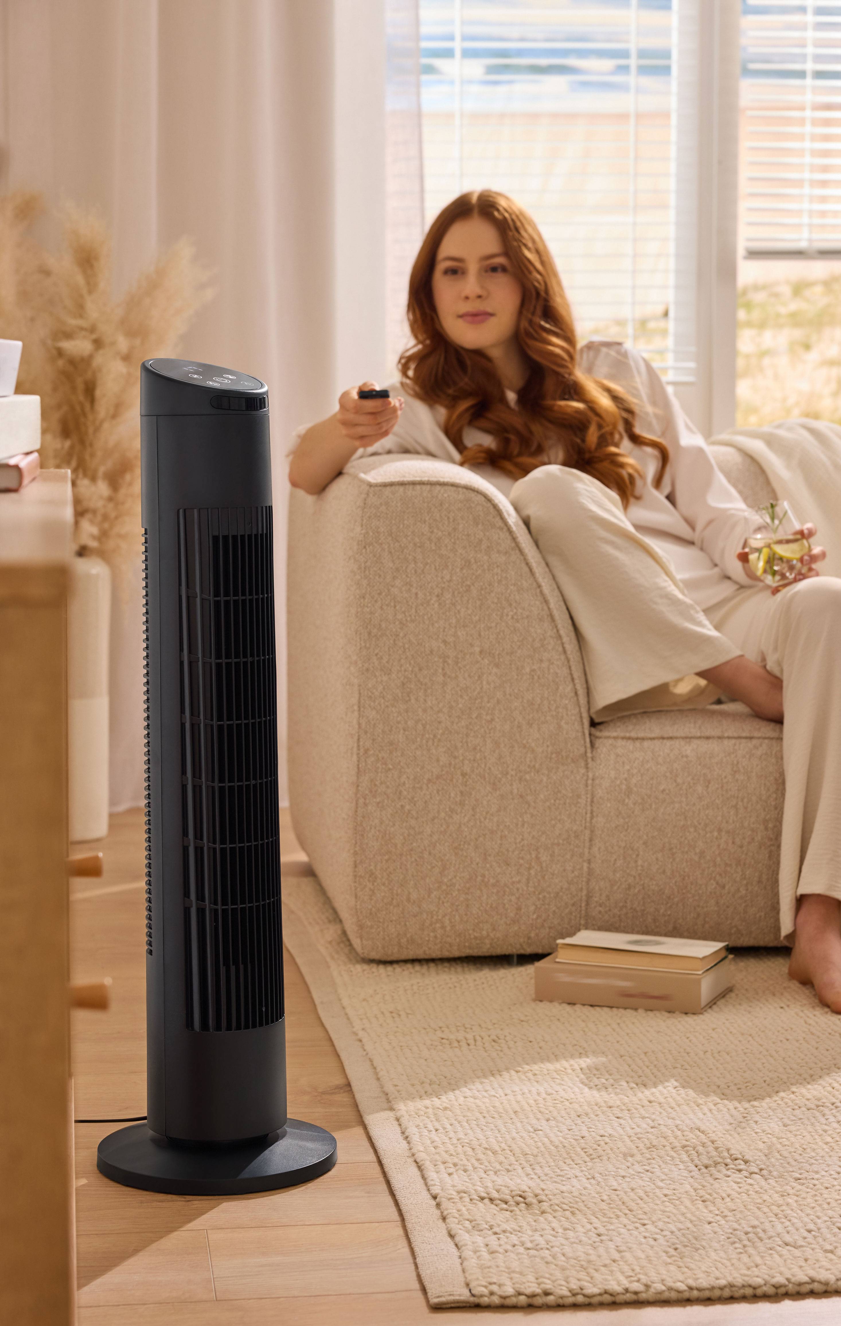 Žena relaxující na pohovce s věžovým ventilátorem v obývacím pokoji.