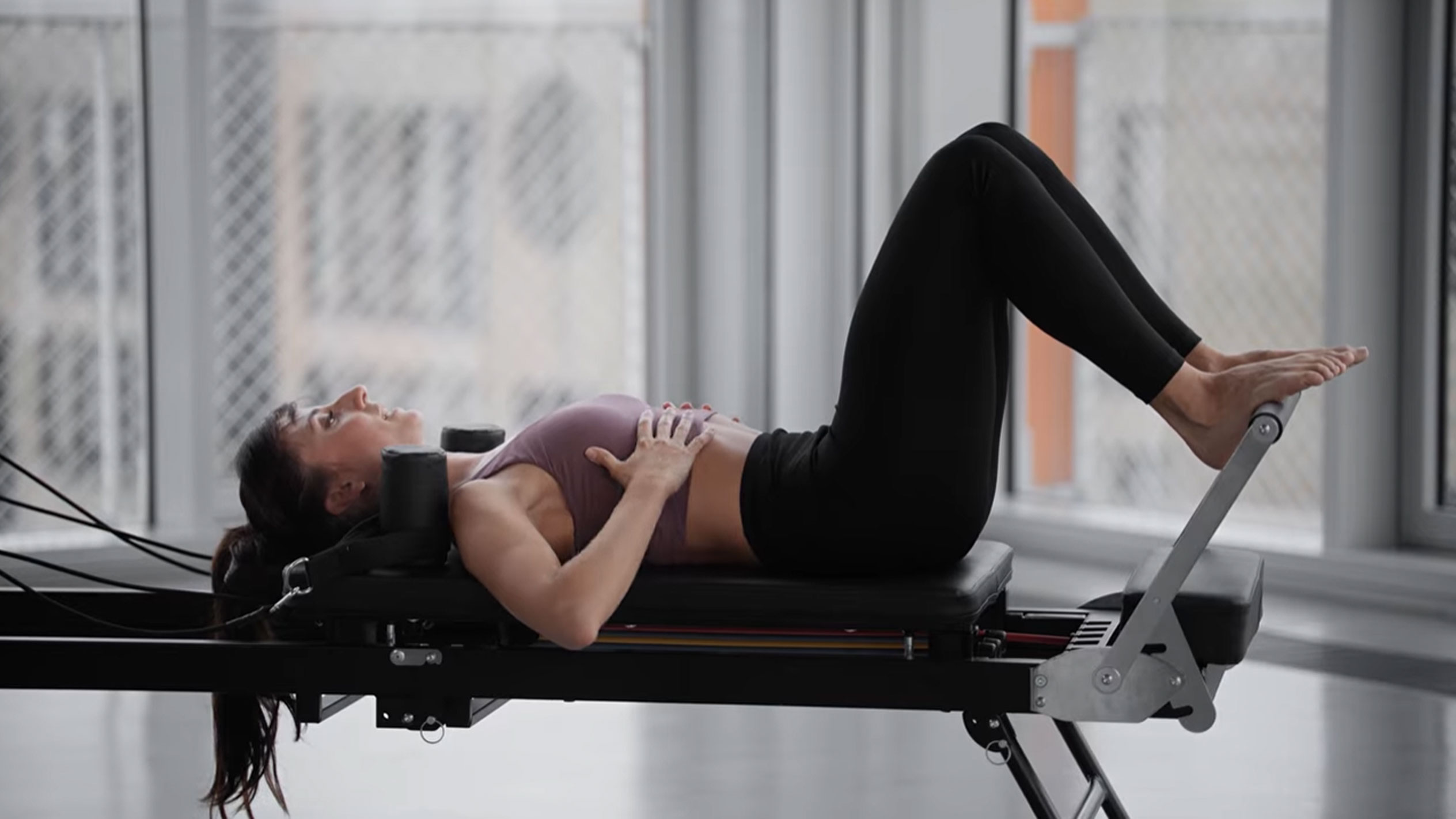 Žena cvičící pilates na reformeru, s rukama na hrudi.
