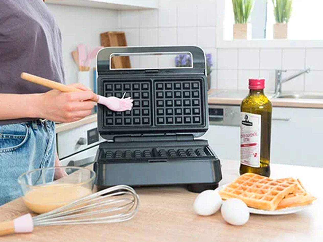 Osoba potírá vaflovač štětcem, s ingrediencemi a hotovými vaflemi na stole.