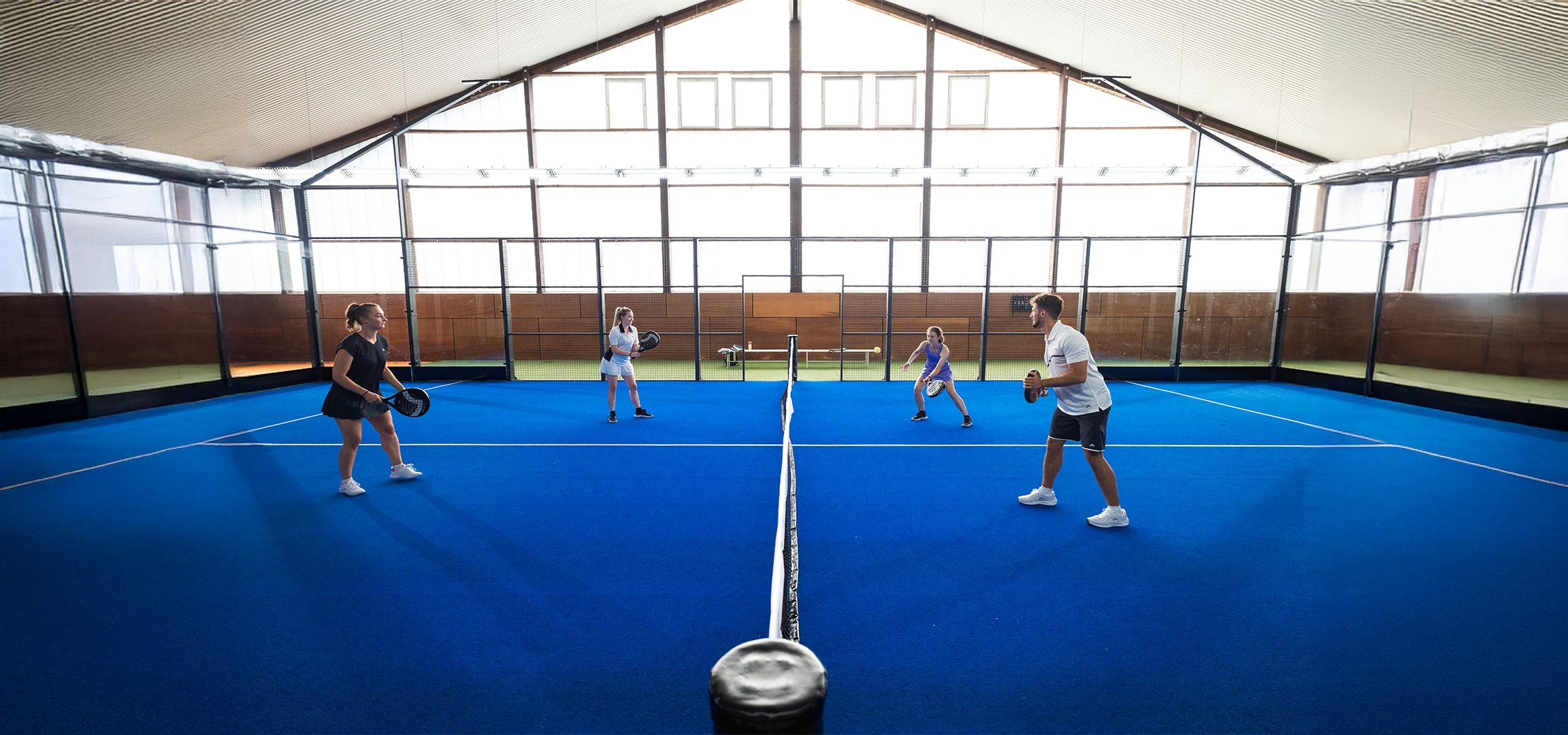 Čtyři lidé hrají padel na modrém kurtu, s raketami a sportovním oblečením.