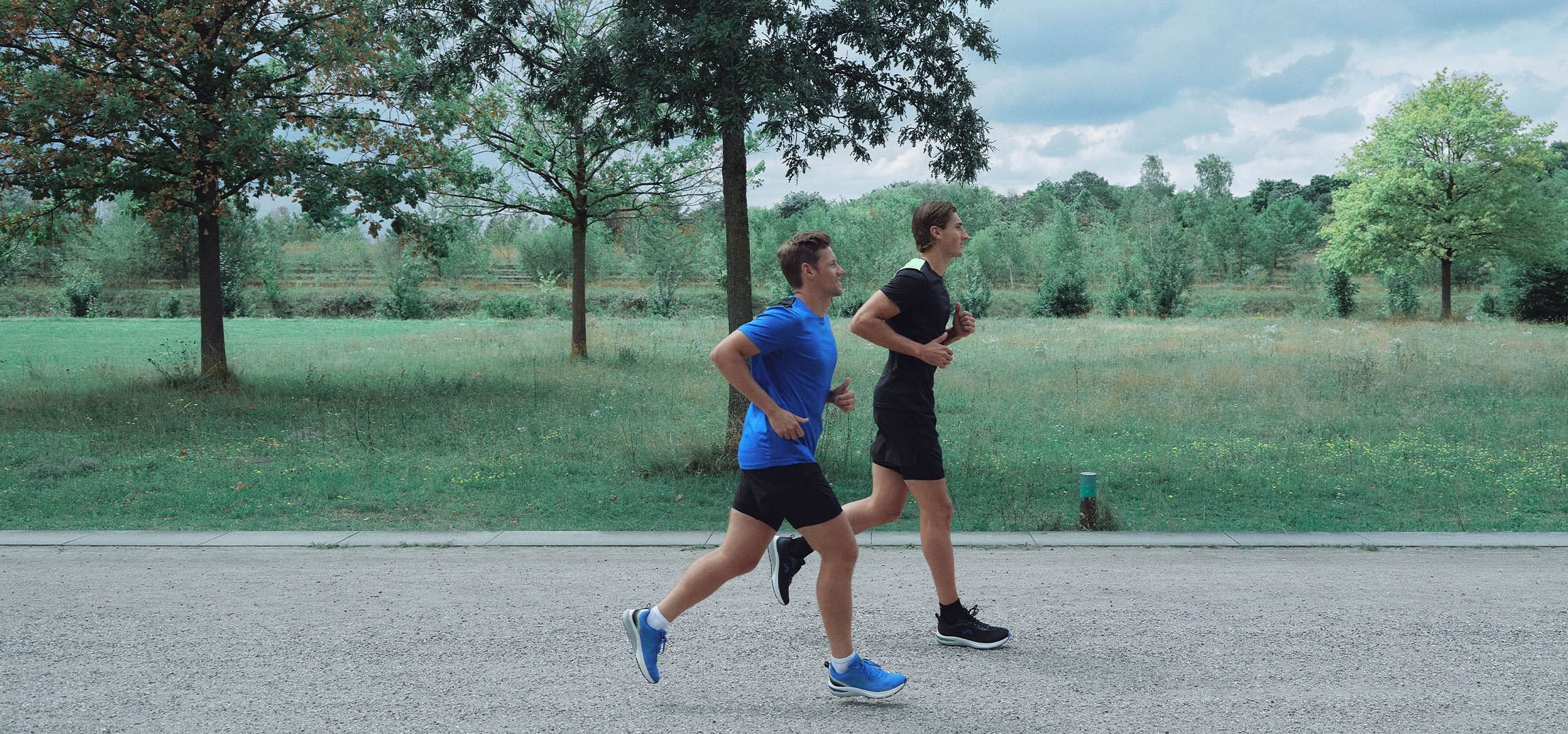 Dva muži běží po štěrkové cestě v parku, oblečeni ve sportovním oblečení.
