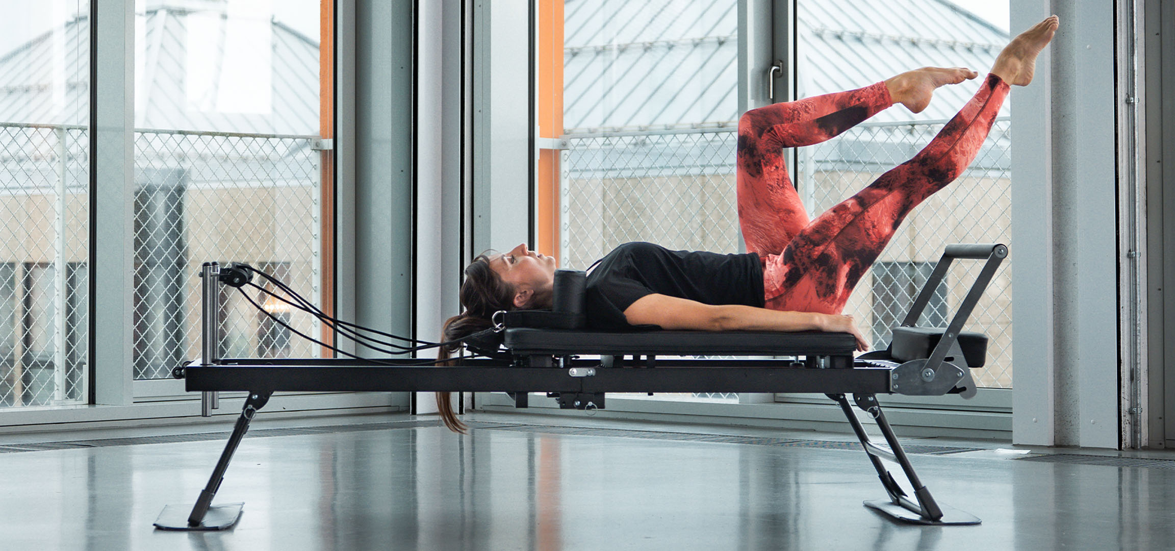 Žena cvičící Pilates na reformer stroji, s fitness doplňky poblíž.