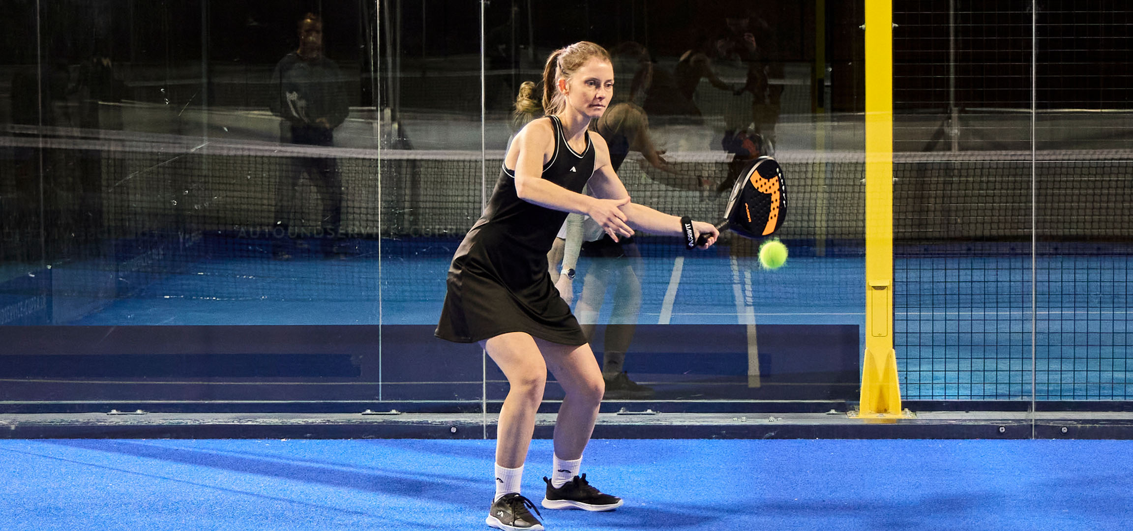 Žena hrající padel na modrém kurtu, oblečená v černém sportovním oblečení.