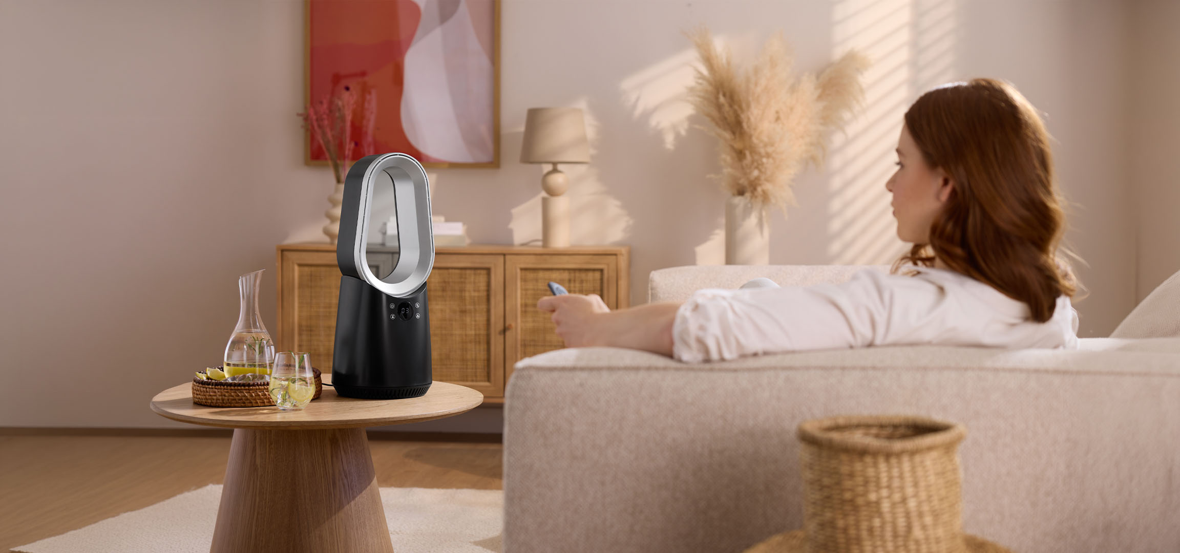 Žena na pohovce s dálkovým ovládáním, moderní ventilátor a nápoje na stole.