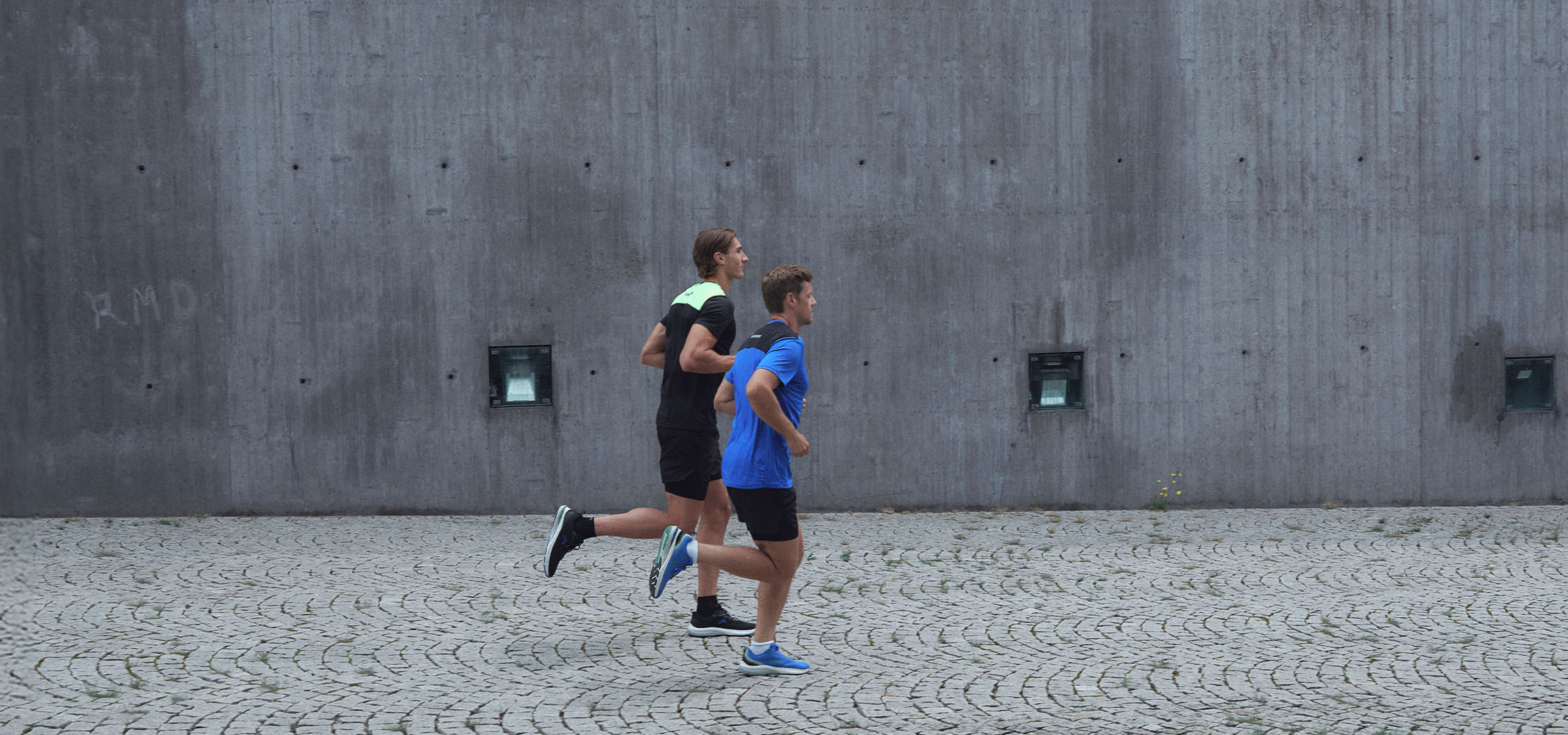 Dva muži ve sportovním oblečení běží podél zdi.