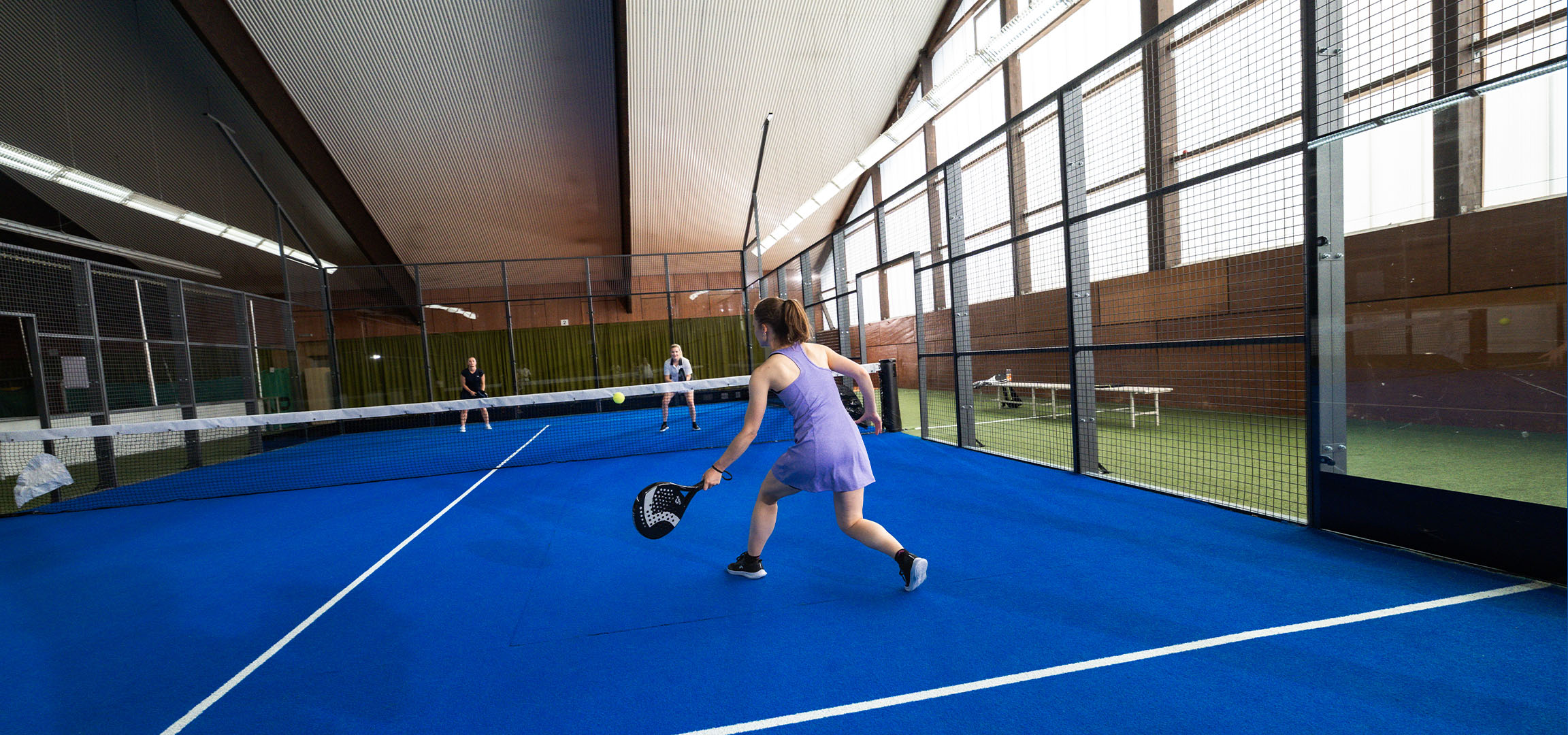 Žena hrající padel na modrém kurtu, s dalšími hráči v pozadí.