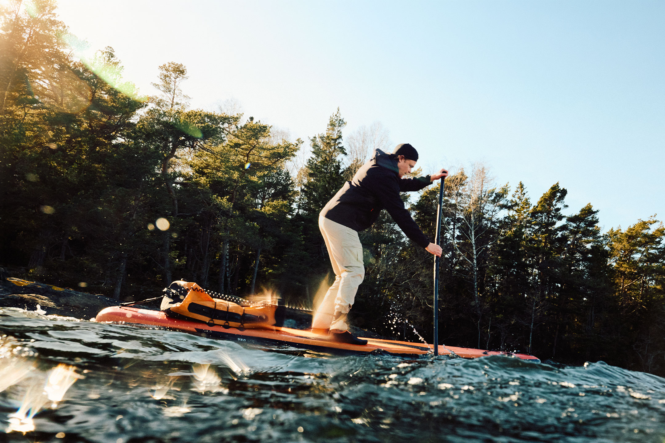 Muž na paddleboardu s oranžovým suchým vakem u lesa.