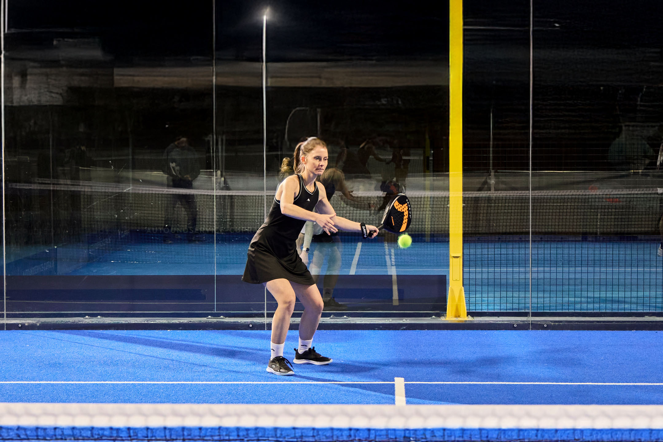 Žena ve sportovním oblečení hraje padel na modrém kurtu s raketou a míčkem.