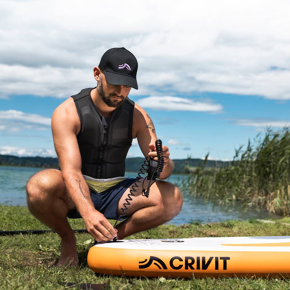 Muž připevňuje bezpečnostní lanko k paddleboardu CRIVIT, v záchranné vestě a kšiltovce.
