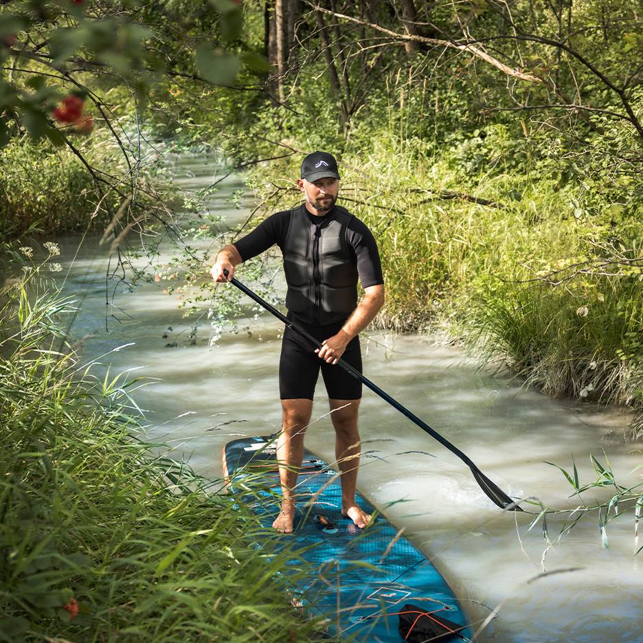 Muž v neoprenu a záchranné vestě stojí na paddleboardu v potoce.