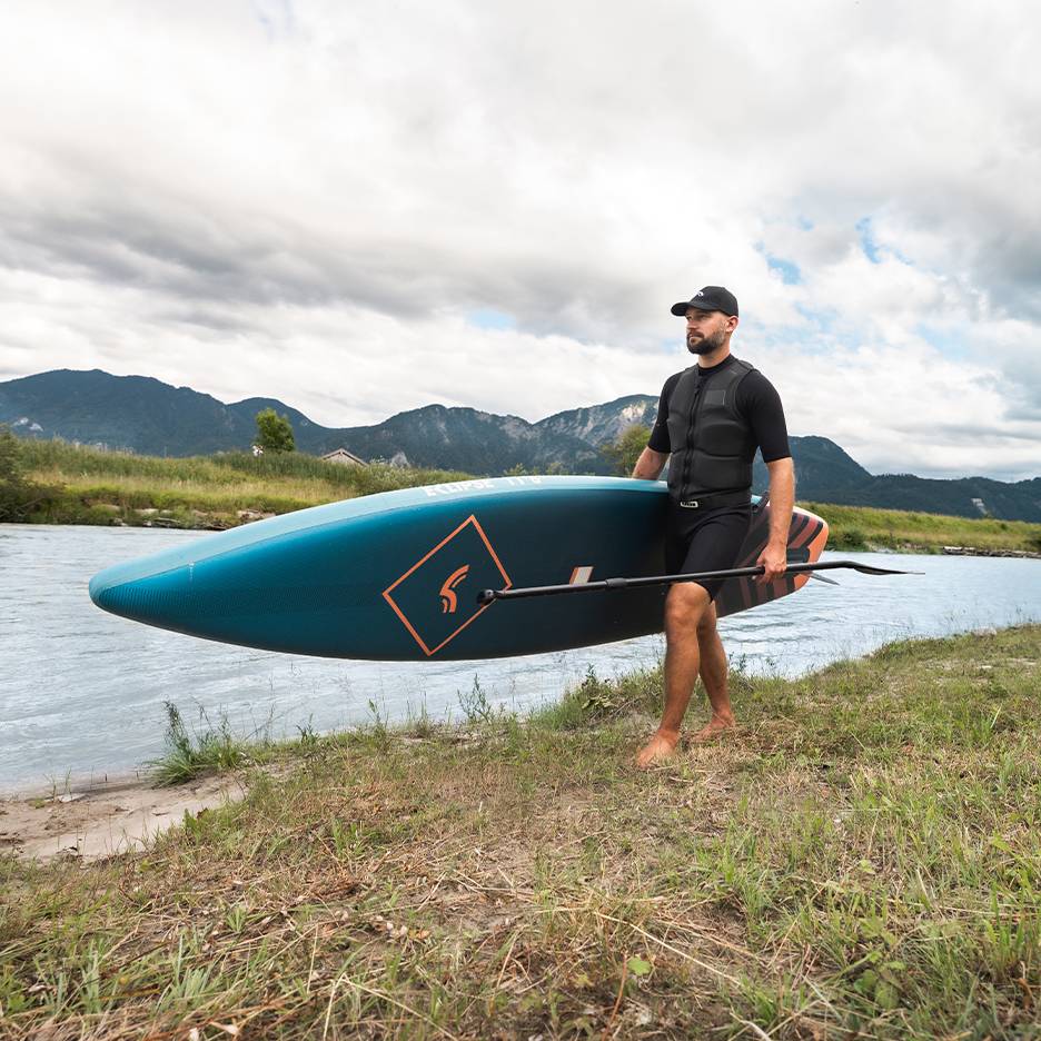 Muž v záchranné vestě a čepici nese paddleboard a pádlo u řeky.