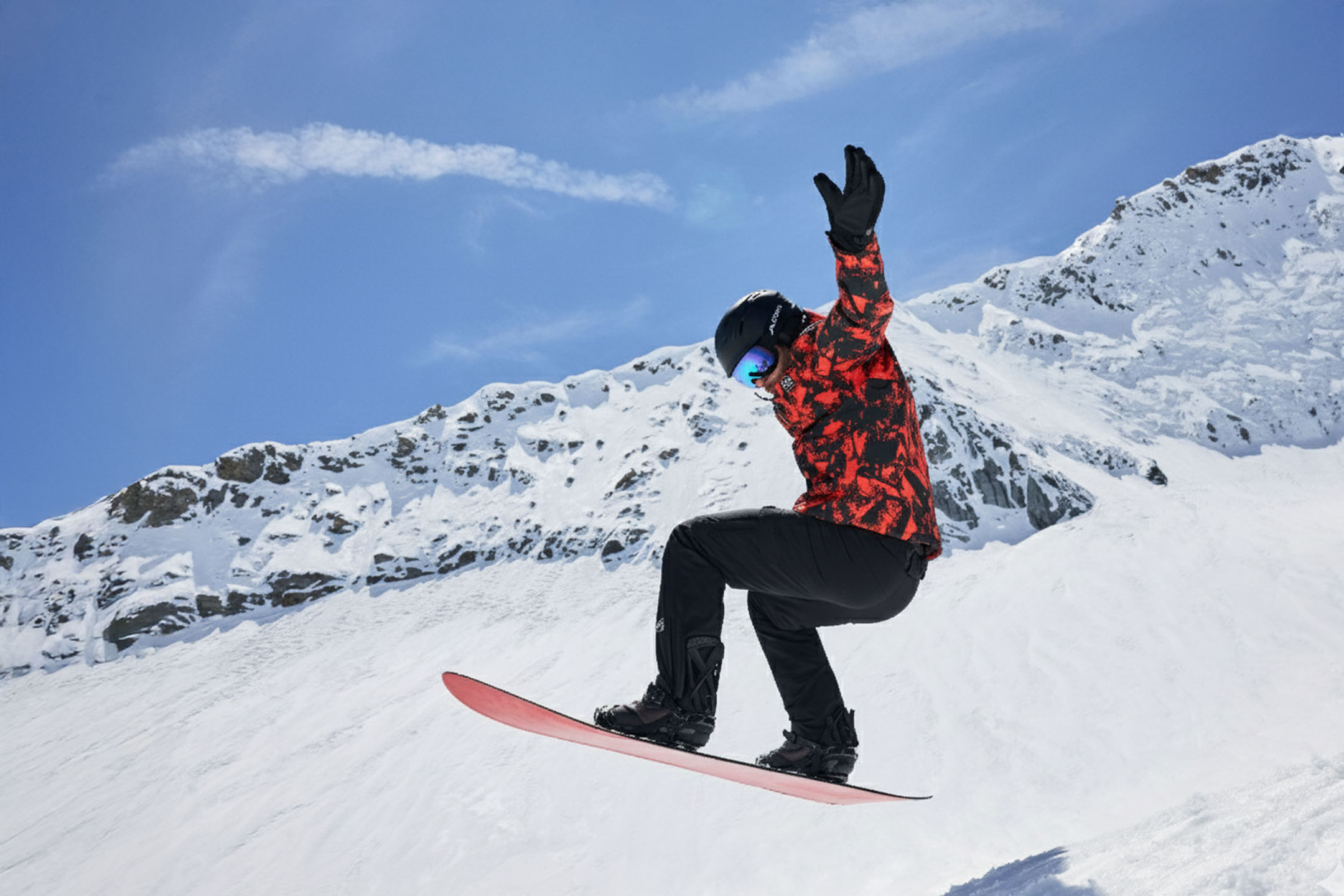 Snowboardista v červeno-černé bundě, černých kalhotách, helmě a brýlích ve vzduchu na zasněžené hoře.
