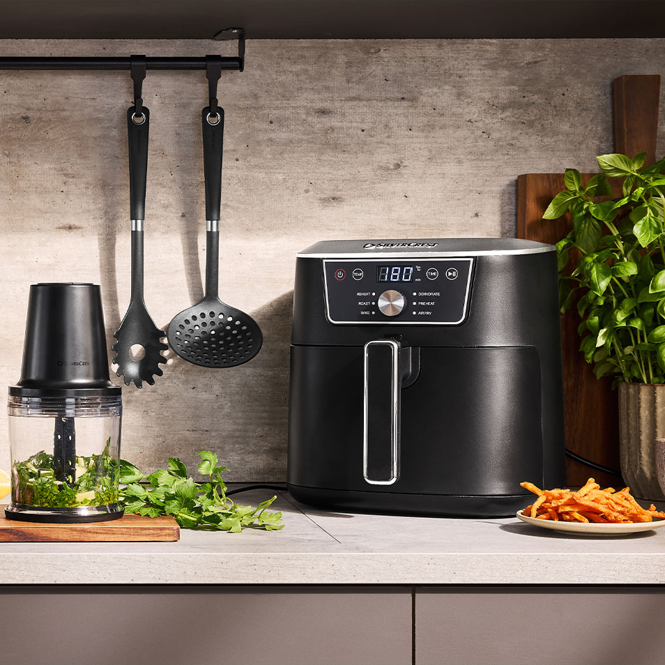 Horkovzdušná fritéza a sekáček SilverCrest na kuchyňské lince s čerstvými bylinkami a hranolky ze sladkých brambor.