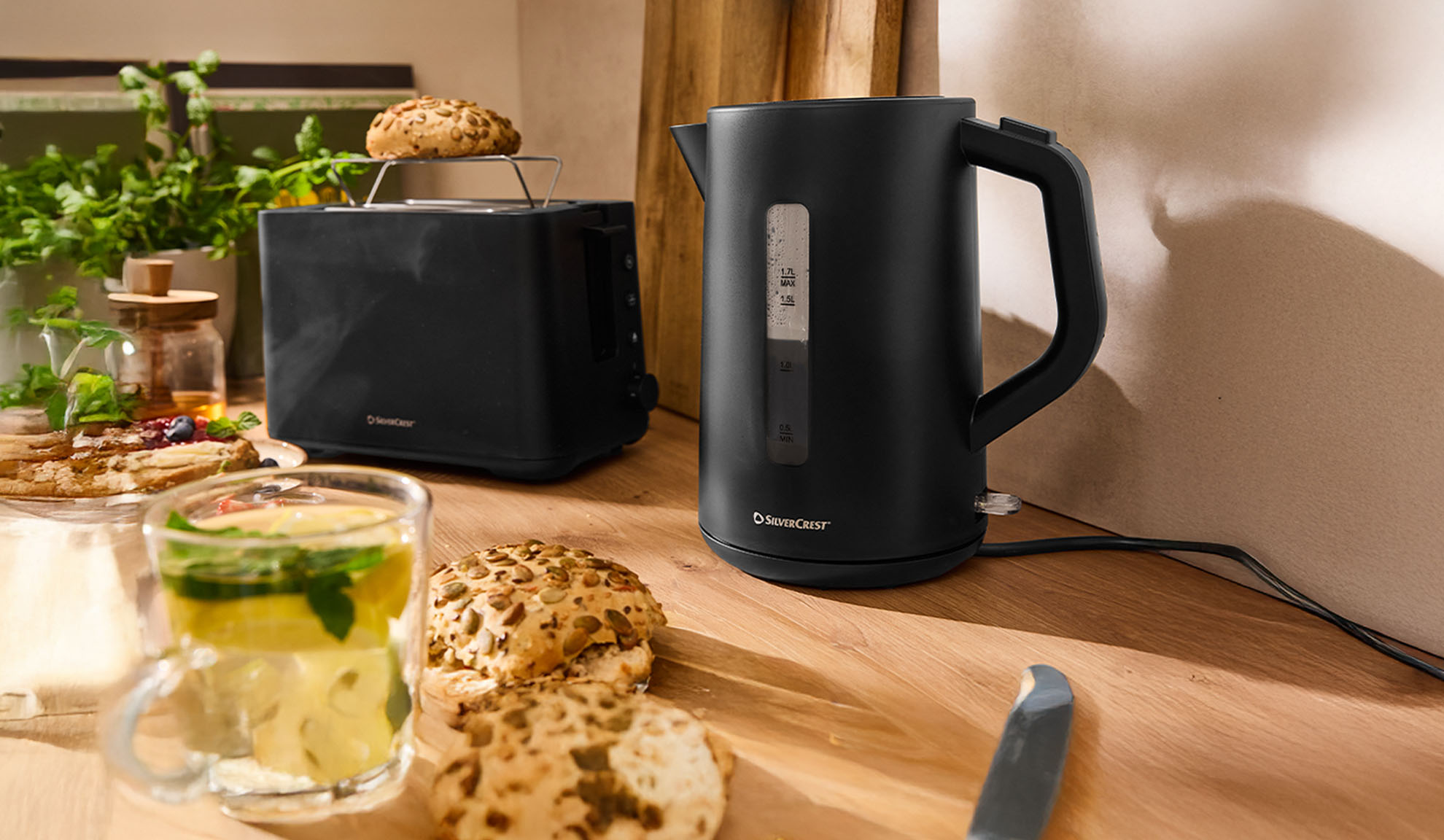 Černá rychlovarná konvice a toustovač SilverCrest na dřevěném stole se snídaní.