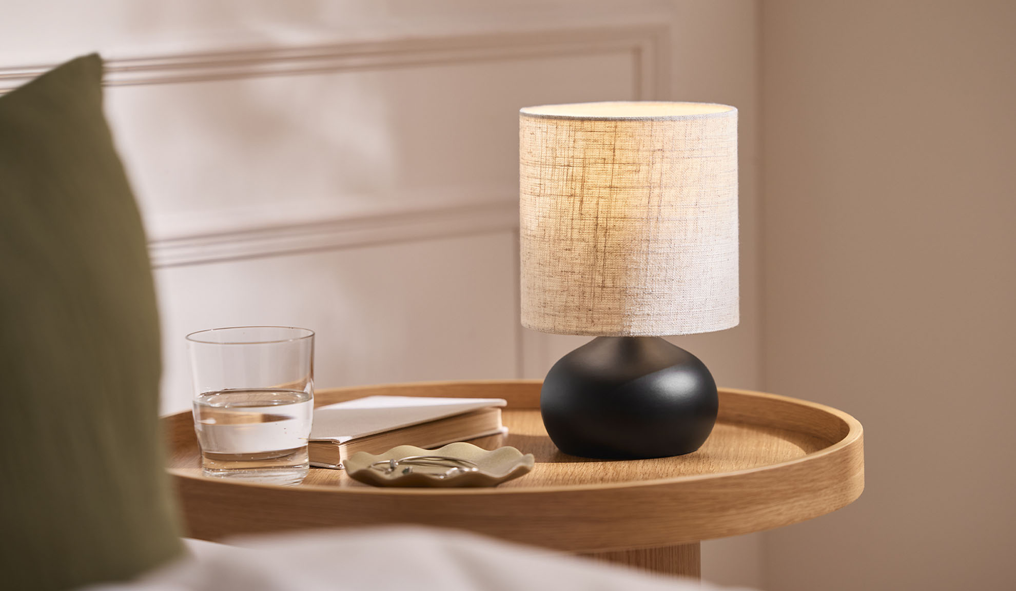 Noční lampa, sklenice, kniha a šperkovnice na dřevěném podnosu vedle postele.