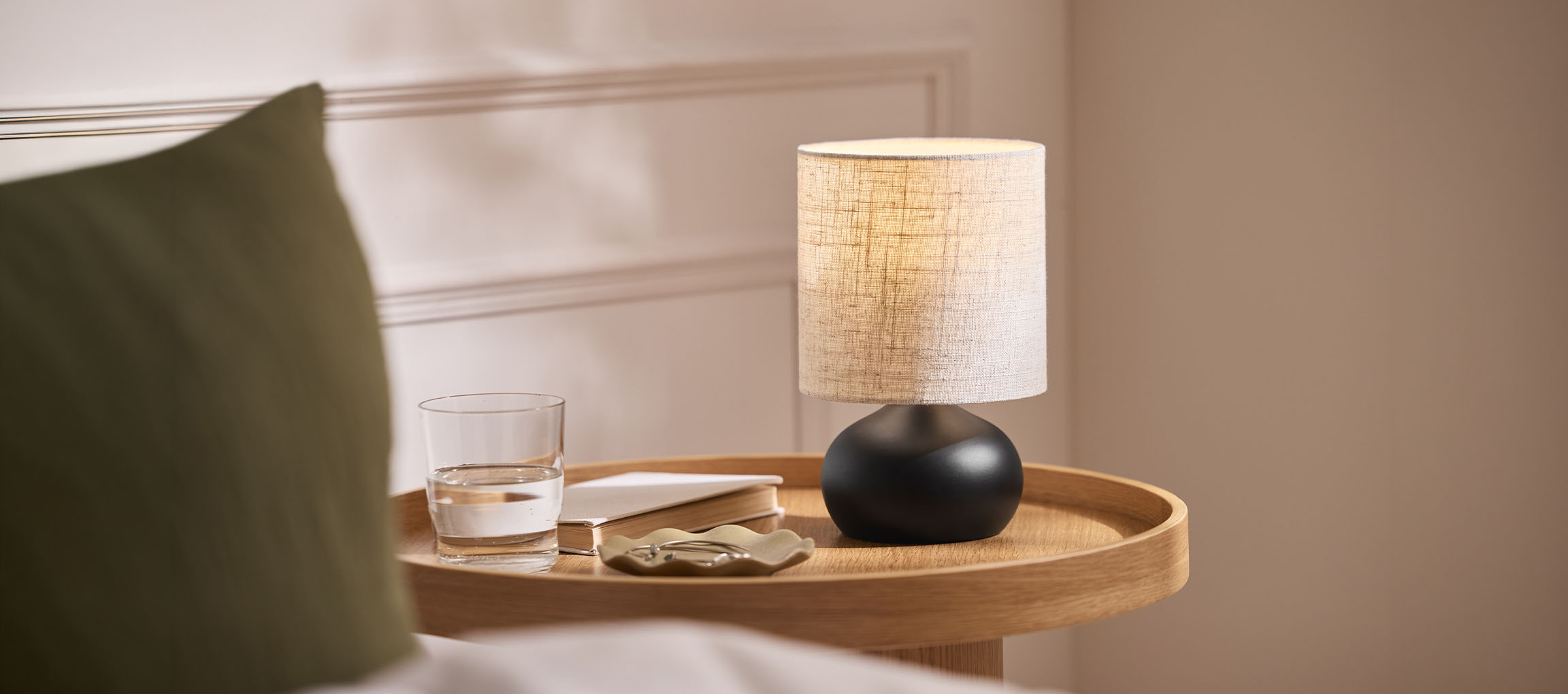 Noční lampa s látkovým stínítkem, sklenice vody a kniha na dřevěném podnosu.