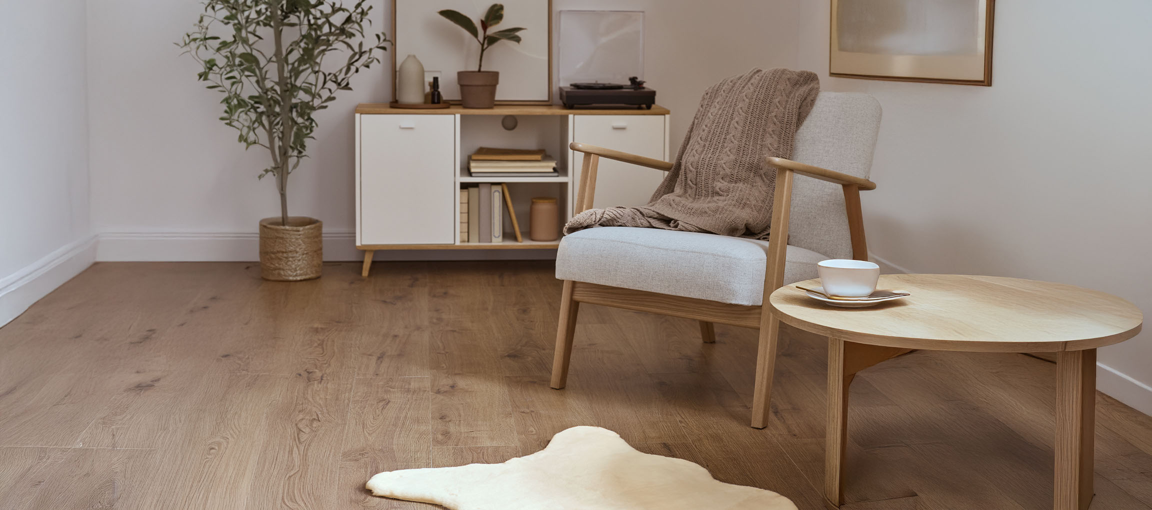 Šedé látkové křeslo s pletenou dekou, dřevěný konferenční stolek a moderní komoda.