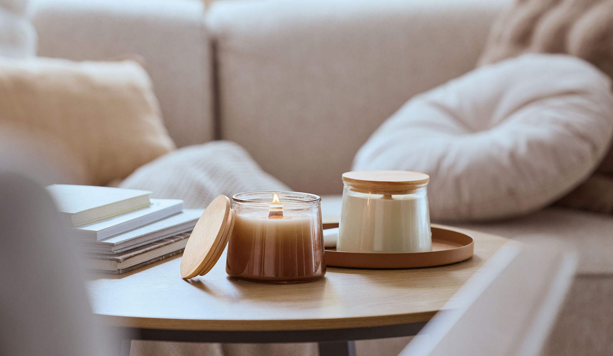 Dvě vonné svíčky na dřevěném stole, jedna zapálená, s útulnou pohovkou v pozadí.