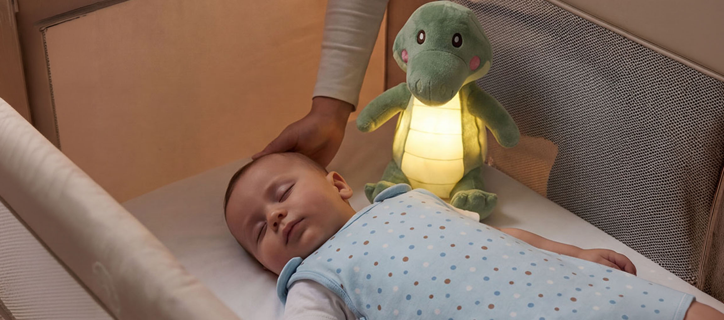 Dítě spí v postýlce s plyšovou noční lampou, dospělý hladí dítě po hlavě.