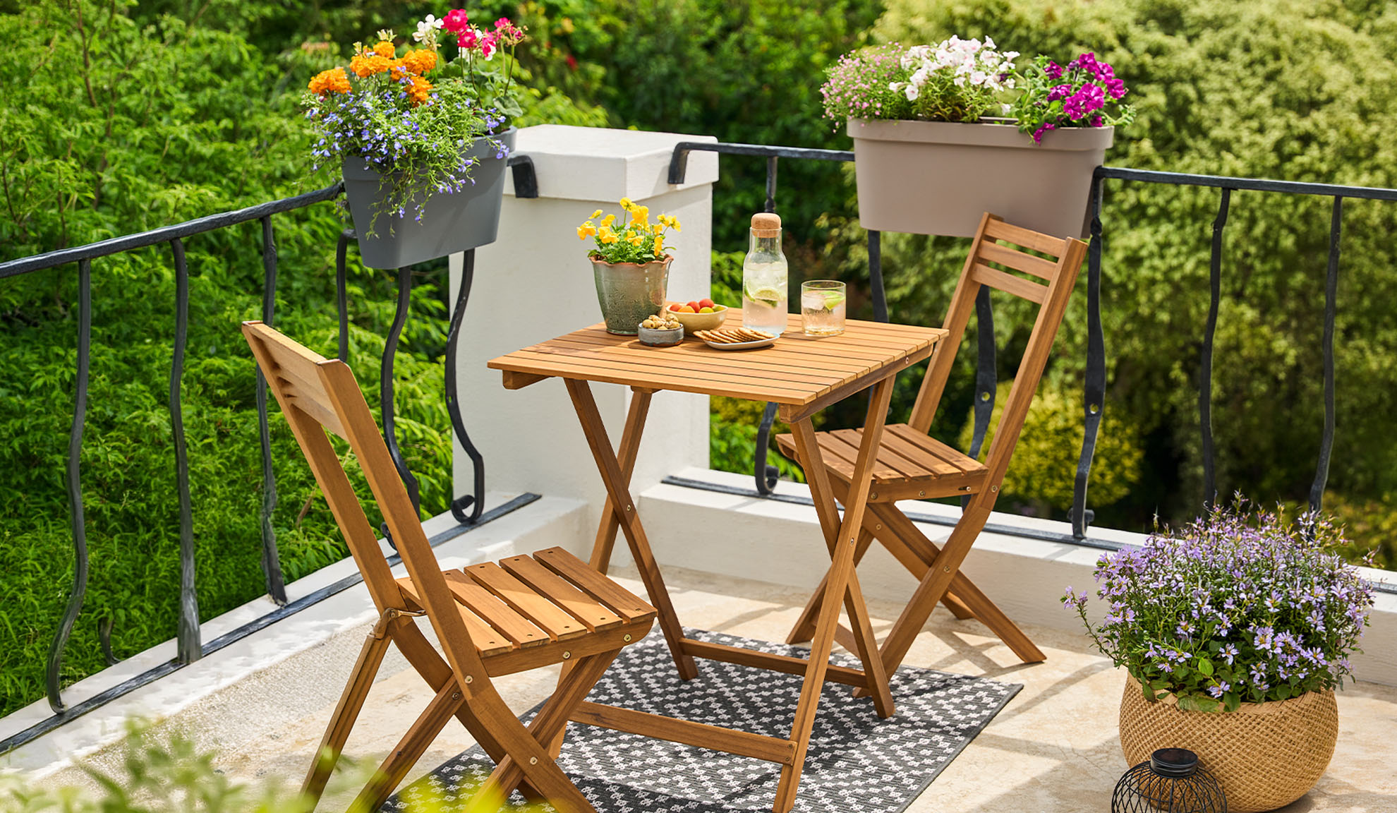 Balkon s dřevěným bistro setem, květinami v květináčích a nápoji na vzorovaném koberci.