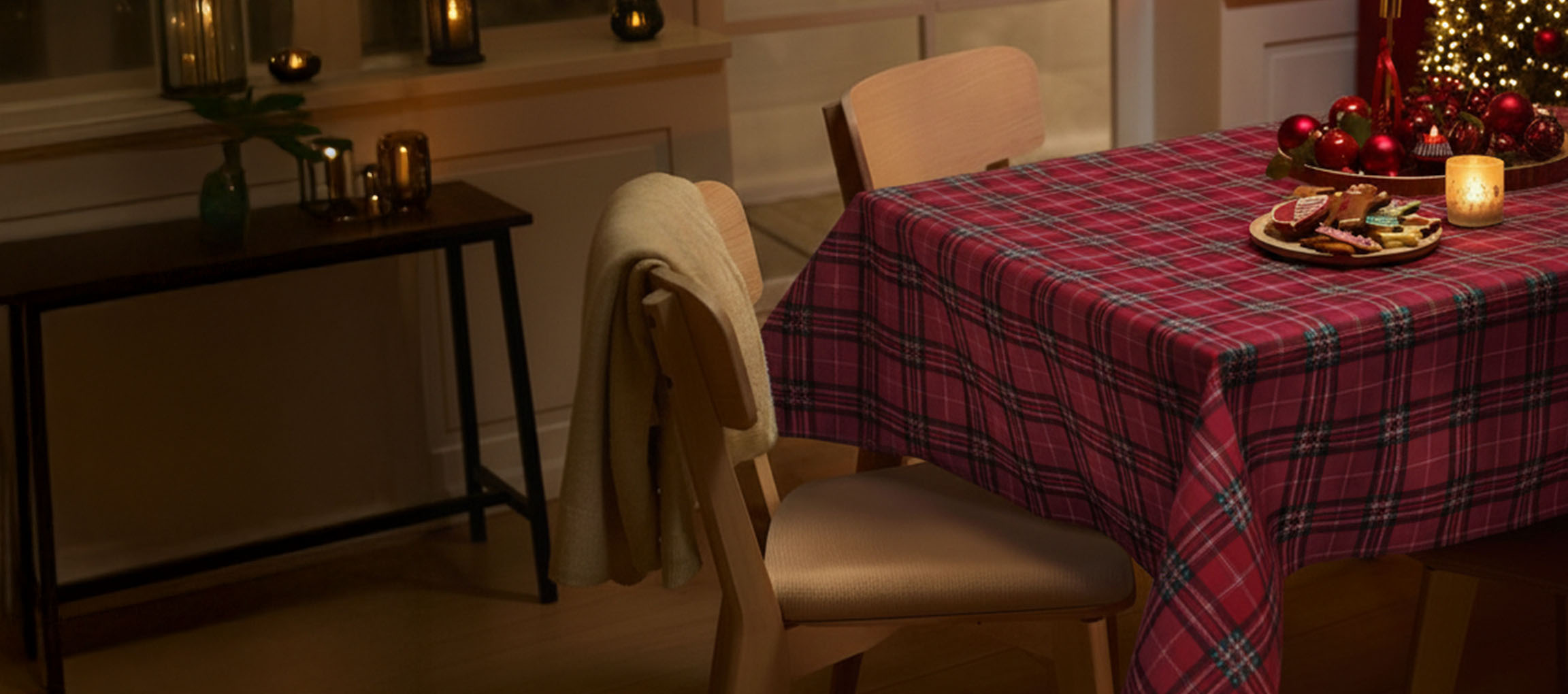 Vánoční stůl s červeným kostkovaným ubrusem, židlemi a vánočním cukrovím.