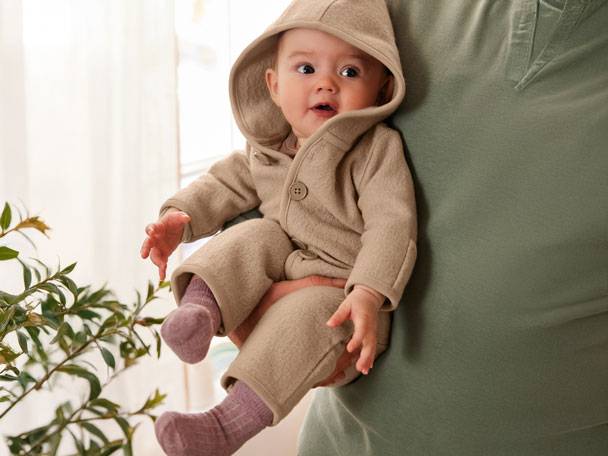 Miminko v béžovém overalu s kapucí, držené dospělým v zeleném tričku.