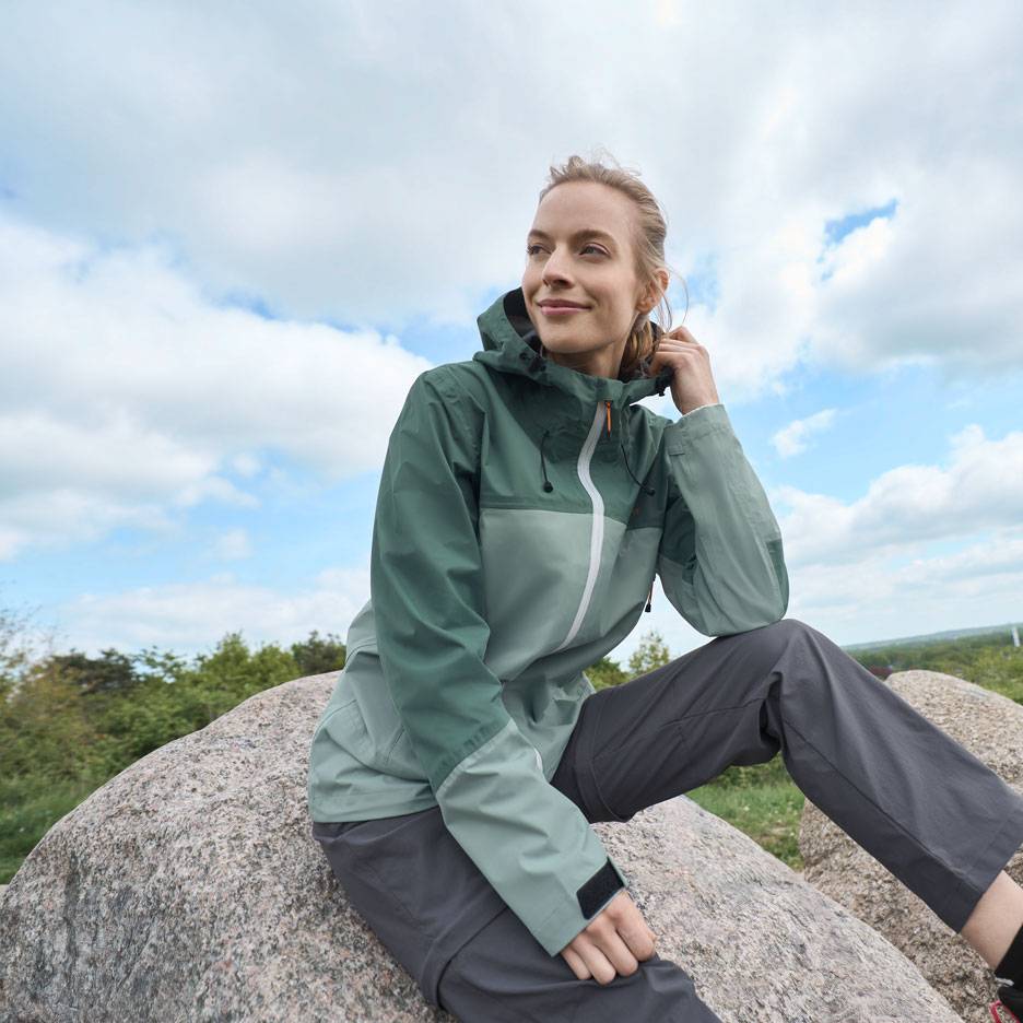 Žena v zelené outdoorové bundě a šedých kalhotách sedí na skále a dívá se do dálky.