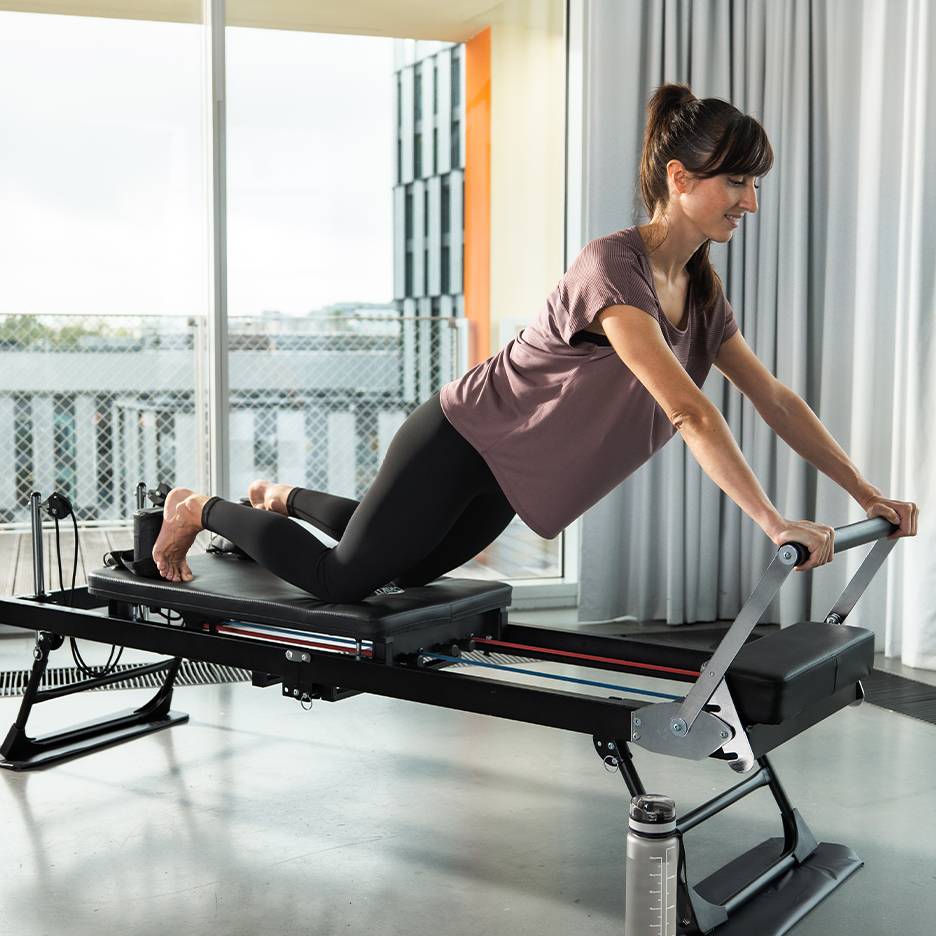 Žena cvičící Pilates na reformer stroji s pružinami a popruhy.