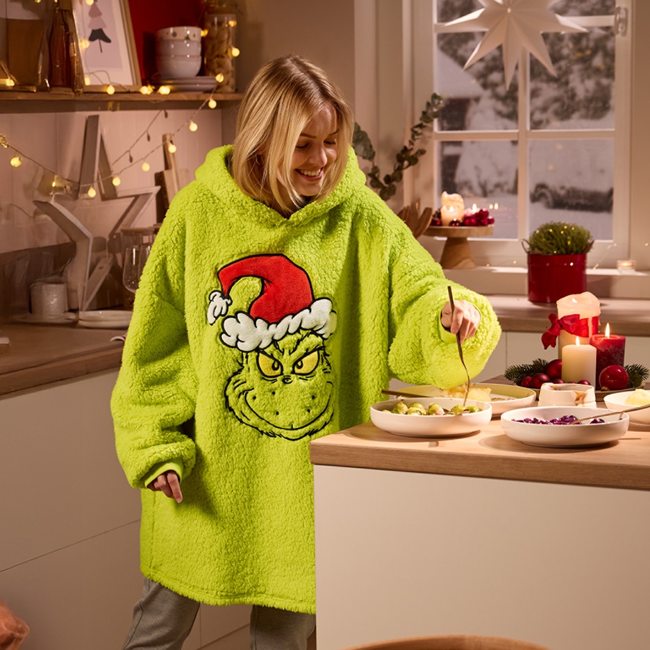 Žena v zelené mikině s motivem Grinche připravuje jídlo v kuchyni s vánoční výzdobou.