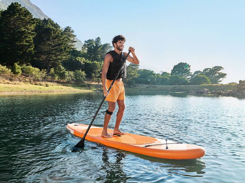 Muž na oranžovém paddleboardu s pádlem na klidné vodě obklopené stromy.