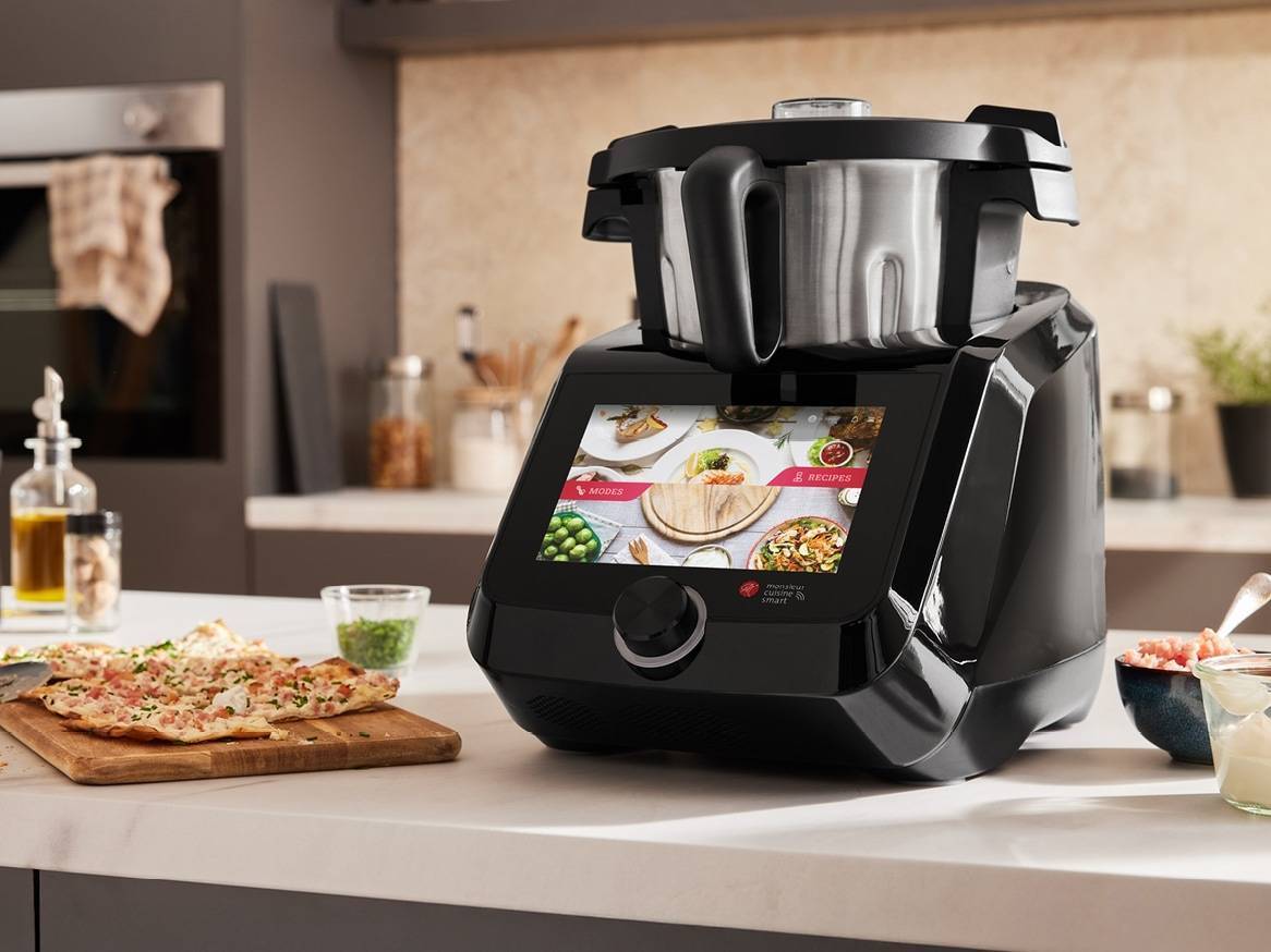 Černý kuchyňský robot s dotykovou obrazovkou zobrazující recepty, na kuchyňské lince s pizzou.