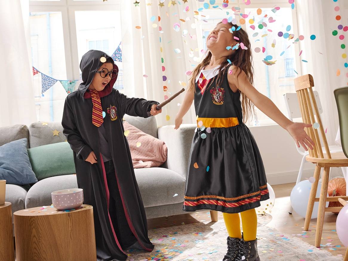 Dvě děti v kostýmech čarodějů si hrají s konfetami v obývacím pokoji.