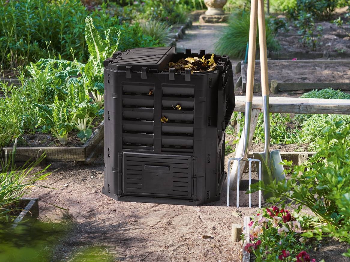 Černý kompostér v zahradě s lopatou a rycími vidlemi.