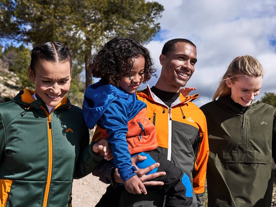 Rodina v barevných outdoorových bundách, muž drží dítě, všichni se usmívají.