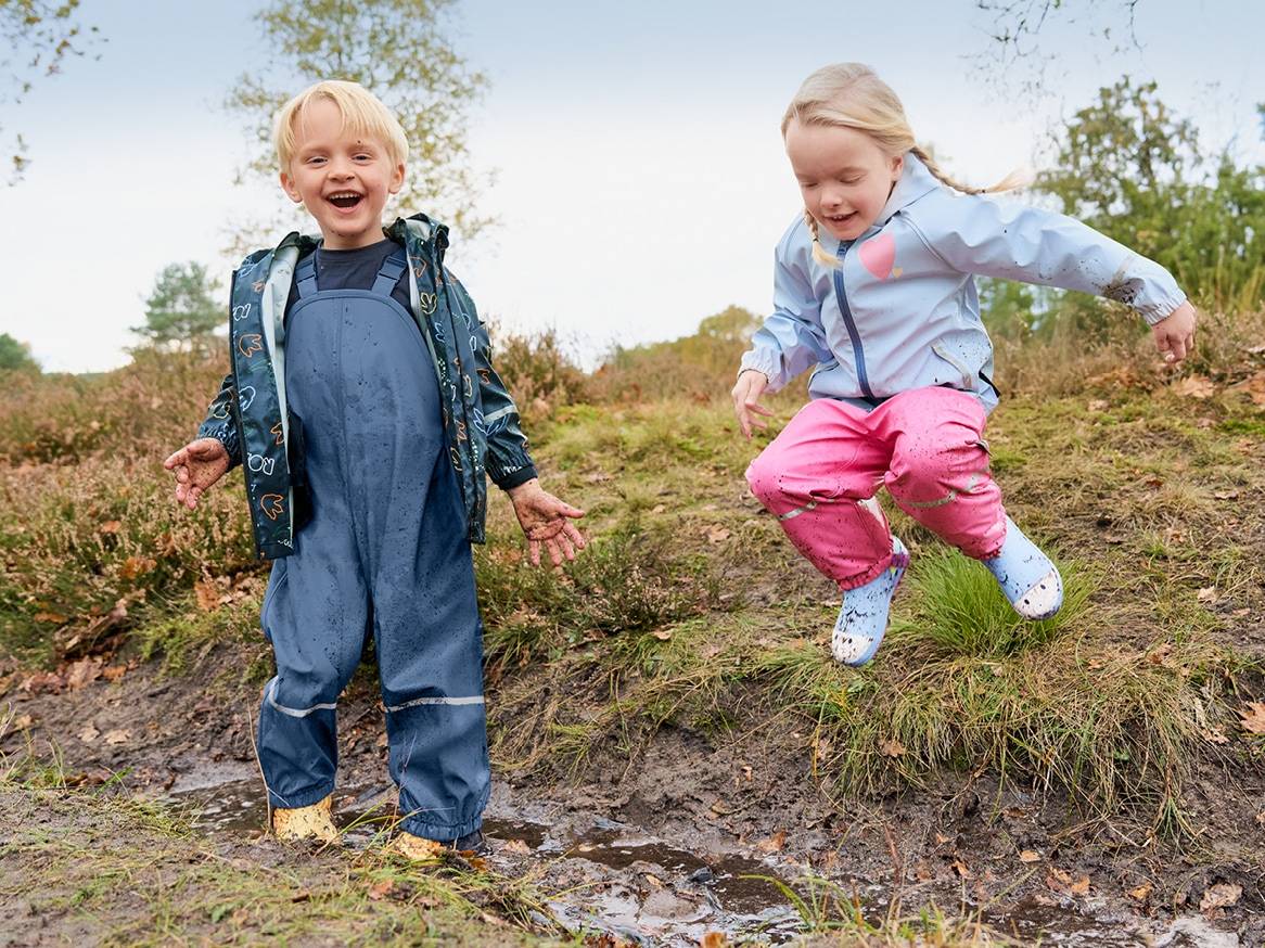 Dvě děti v nepromokavém oblečení a holínkách si hrají v blátě.