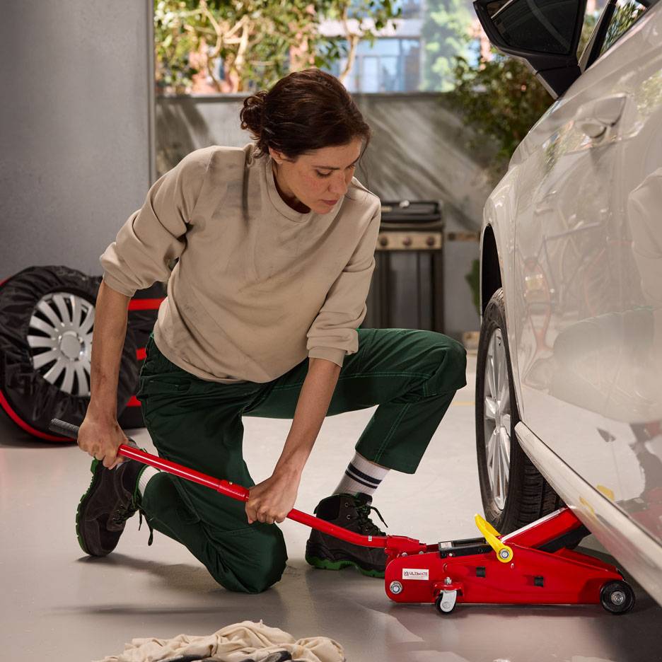 Žena používá červený hydraulický zvedák k zvedání bílého auta, s rezervním kolem poblíž.