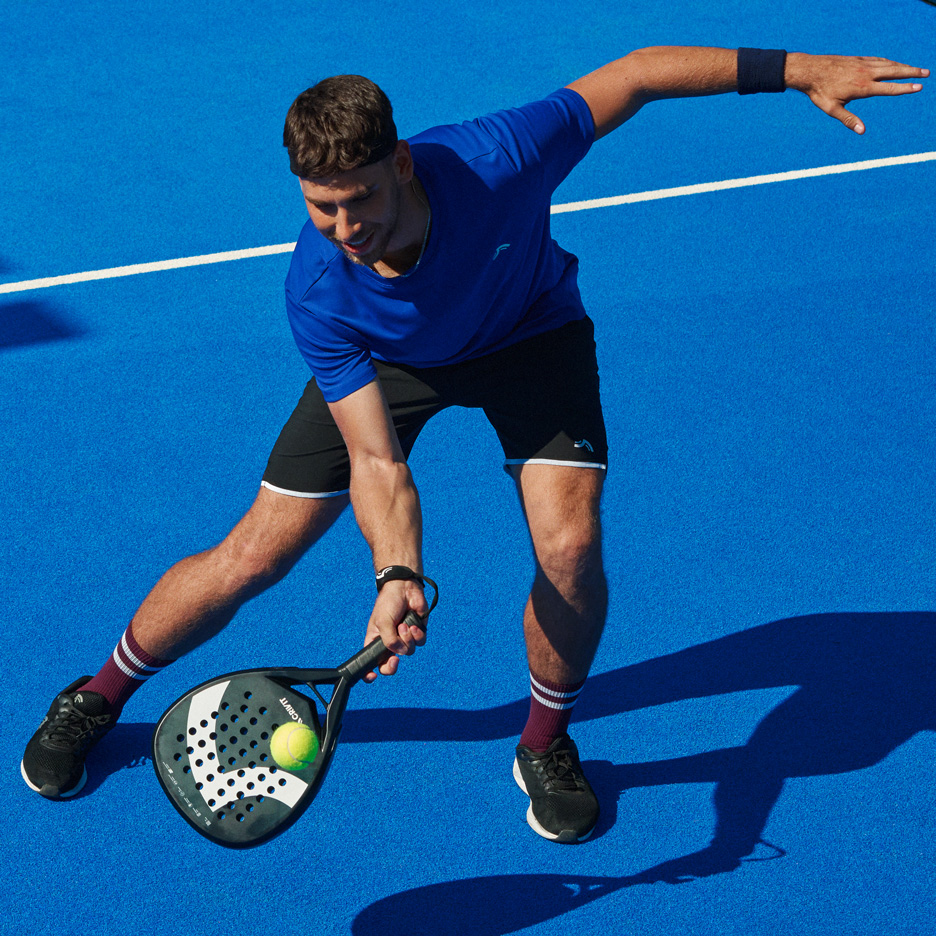 Muž v modrém tričku a černých šortkách hraje padel s raketou a míčkem.