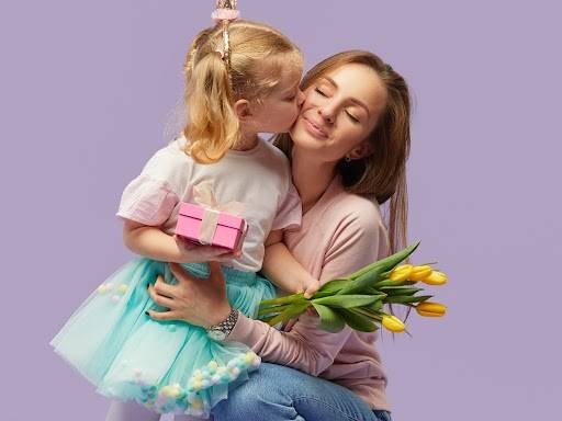 Dívka dává mamince pusu na tvář, drží dárek a maminka drží kytici žlutých tulipánů.